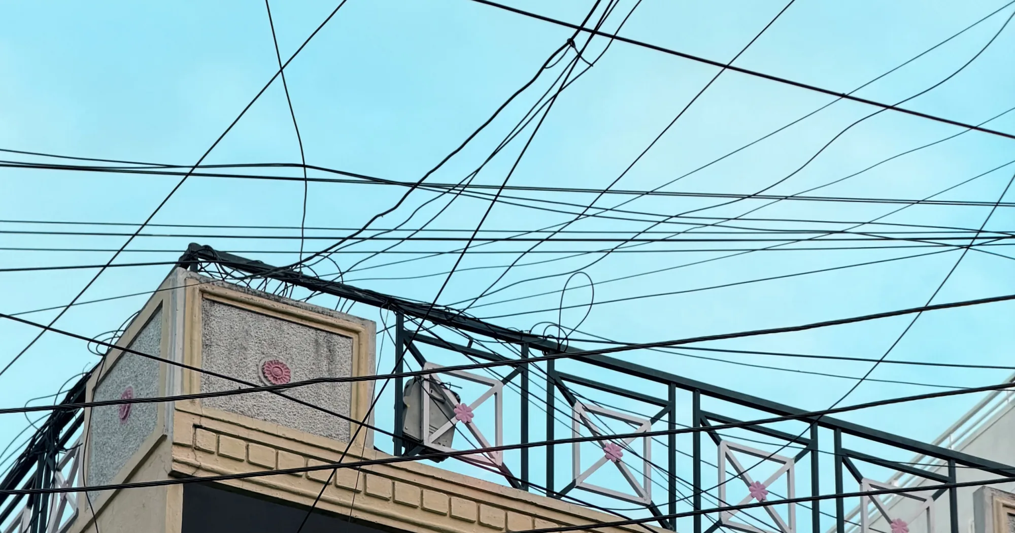 A dense tangle of overhead electrical and telecom wires on a streetside pole, crossing at random angles, sagging in loops, with no discernible order between the cables